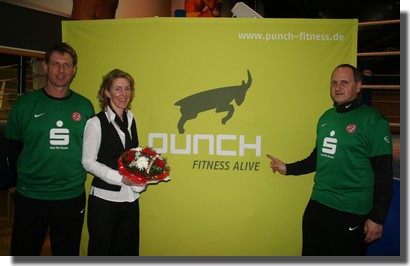 Das RWE-Trainergespann Uwe Erkenbrecher (l.) und Ralf (r.) Aussem überreichen der Punch-Geschäftsführerin Rebekka Bisten einen rot-weissen Blumenstrauß